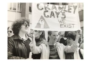 Black and white image of woman holding up a sign which says "Crawley Gays, We Exist"